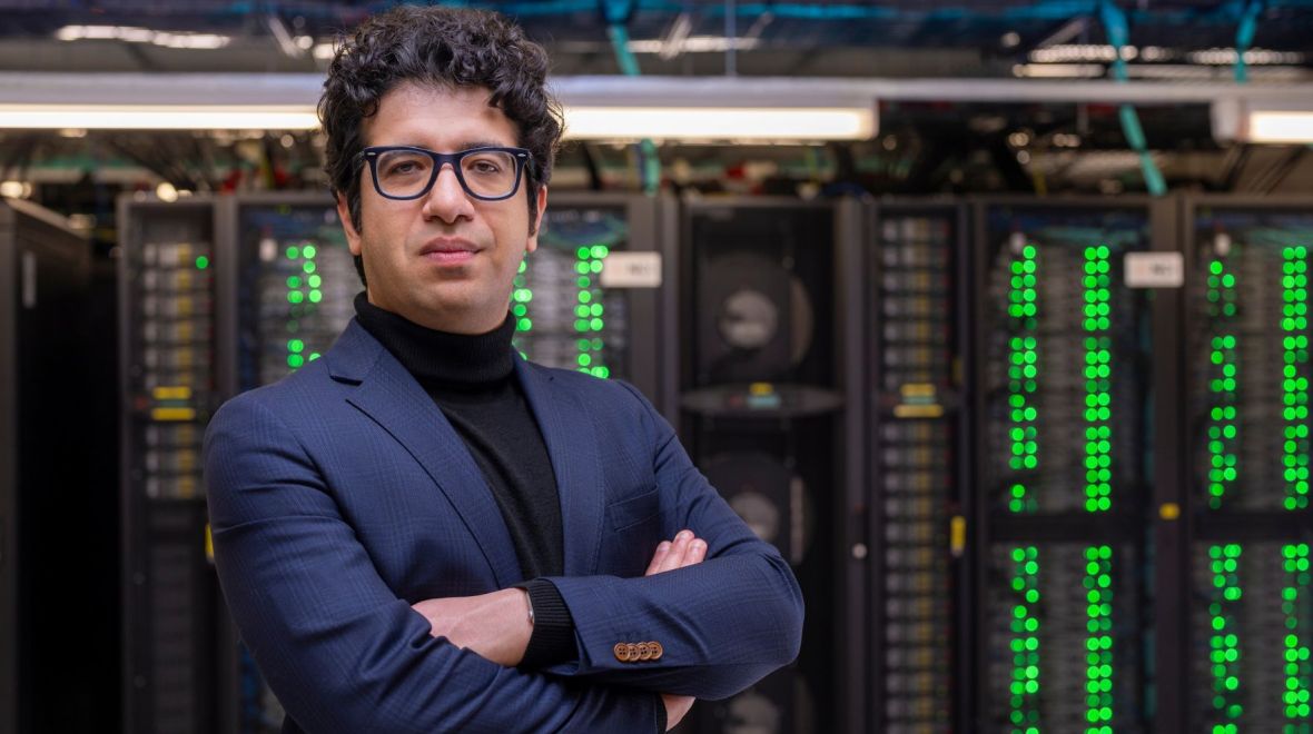 Dr Eli Niktab stands with arms folded in front of computer server racks with glowing green lights.